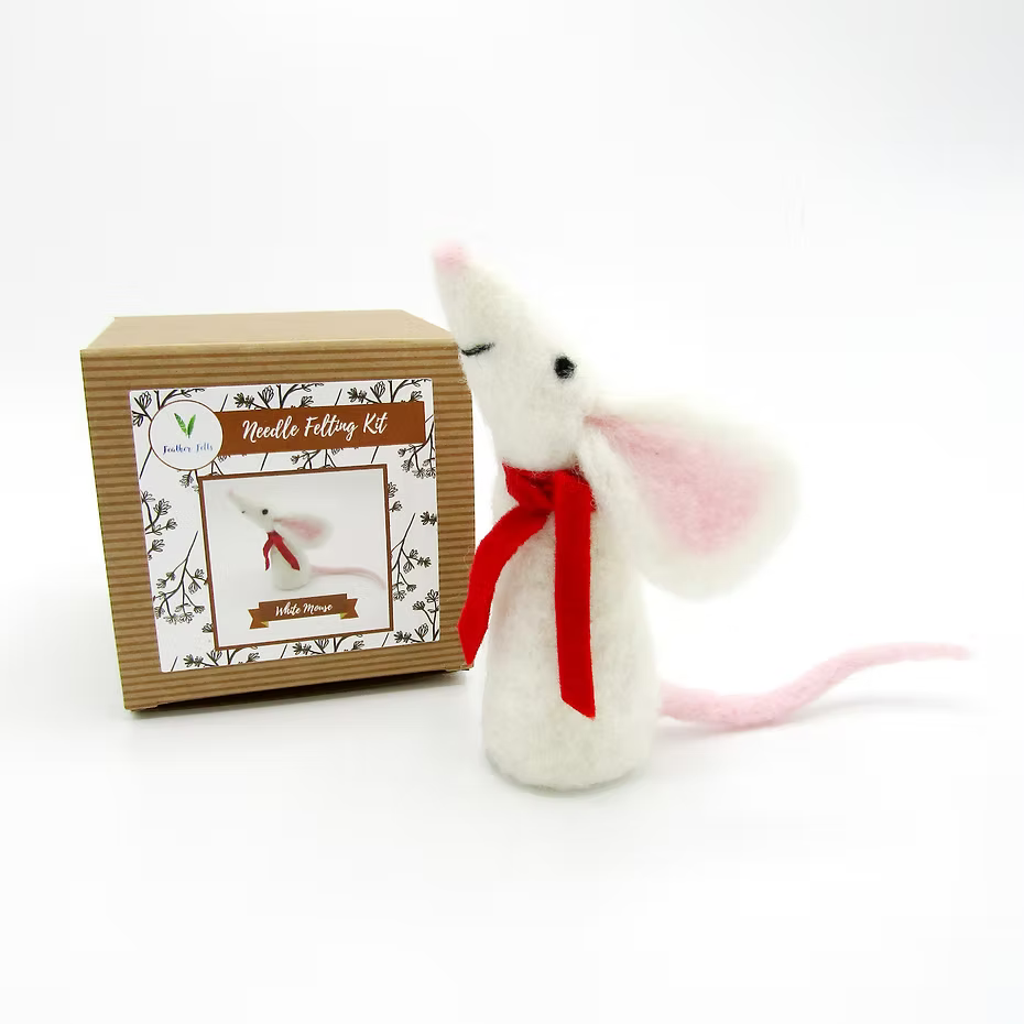 A felted white mouse with pink ears and tail, wearing a red scarf, stands beside a Feather Felts White Mouse - Needle Felting Kit No Foam box, which shows the mouse and includes needle felting instructions on the front.