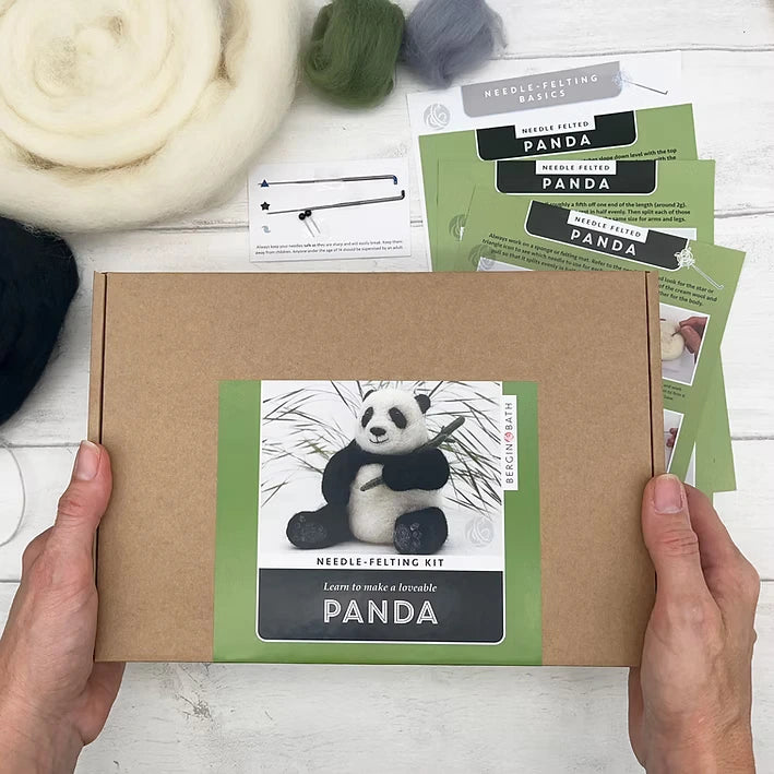 A person holds a Panda Needle Felting Kit box featuring a felted panda photo. Around it are wool roving, felting needles, and instructions—everything beginners need for needle felting—all on a white surface.