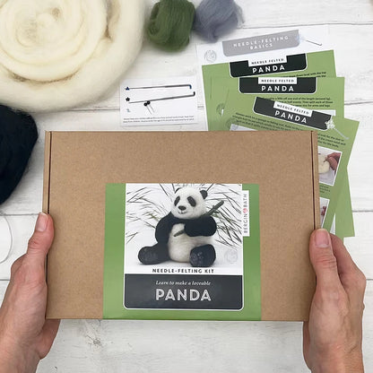 A person holds a Panda Needle Felting Kit box featuring a felted panda photo. Around it are wool roving, felting needles, and instructions—everything beginners need for needle felting—all on a white surface.