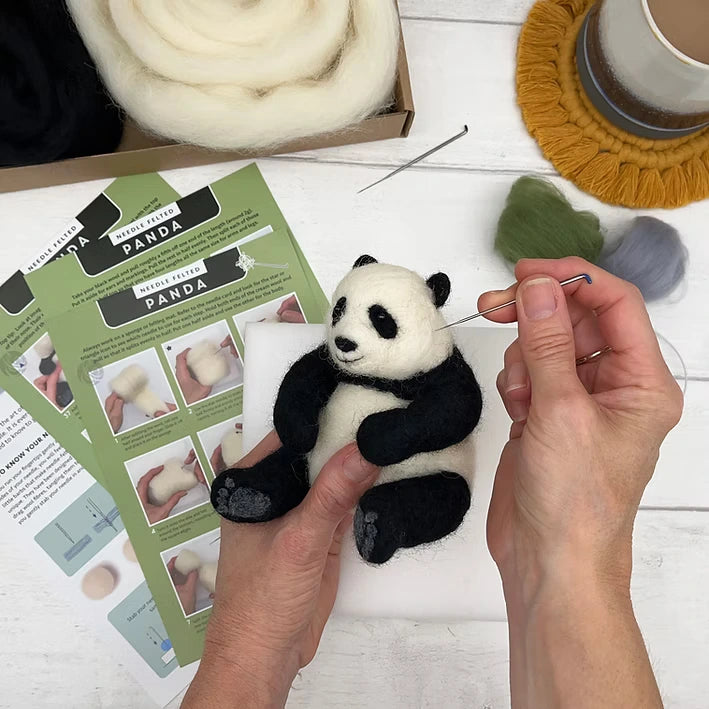 Using the Panda Needle Felting Kit, a person crafts a small wool panda bear. The kit includes felting needles, instructions, and wool roving—ideal for beginners. A cup on a yellow coaster sits nearby for added comfort while creating.