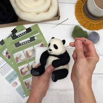 Using the Panda Needle Felting Kit, a person crafts a small wool panda bear. The kit includes felting needles, instructions, and wool roving—ideal for beginners. A cup on a yellow coaster sits nearby for added comfort while creating.