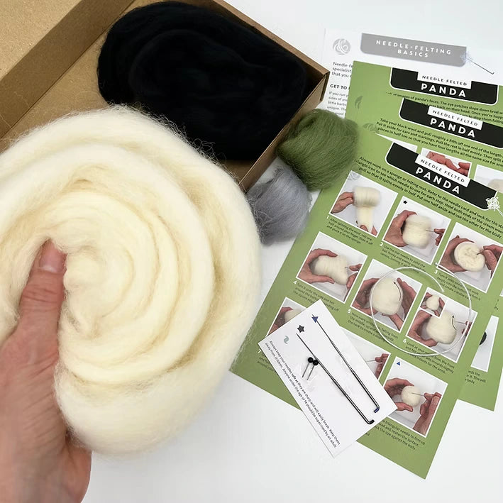 A hand holds white wool roving next to the Panda Needle Felting Kit, which comes with black, green, and gray wool, felting needles, and illustrated instructions—ideal for beginner needle felting.