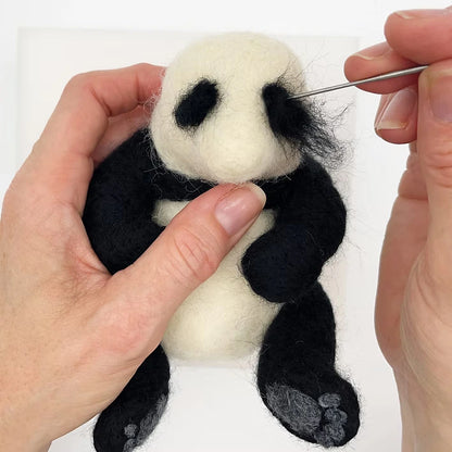 Using the Panda Needle Felting Kit, a person adds black wool details to a small felt panda's face with a needle, gently holding the panda—ideal for beginners exploring needle felting.
