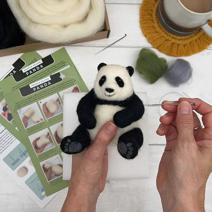 Using the Panda Needle Felting Kit, a person needle felts a small wool panda on a foam pad. The kit includes instructions, wool roving, and tools—all shown on a white table with a mug on a yellow coaster in the background.