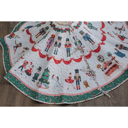 The Nutcracker Tradition Tree Skirt Kit by Quilted Joy features Riley Blake Nutcracker fabric with festive illustrations, quilted design, green and red borders, and is shown on a wooden floor.