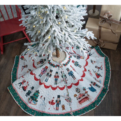 A white pre-lit Christmas tree stands on the Nutcracker Tradition Tree Skirt Kit by Quilted Joy, which displays colorful Nutcracker-themed designs. Wrapped gifts and a red chair sit nearby on a dark wood floor.