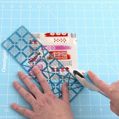 Using the Omnigrid 12 x 18 Compact Rotary Cutting Kit by Tacony Corporation, a person cuts patterned fabric on a blue, measured mat with the rotary cutter and clear grid ruler included in the kit.