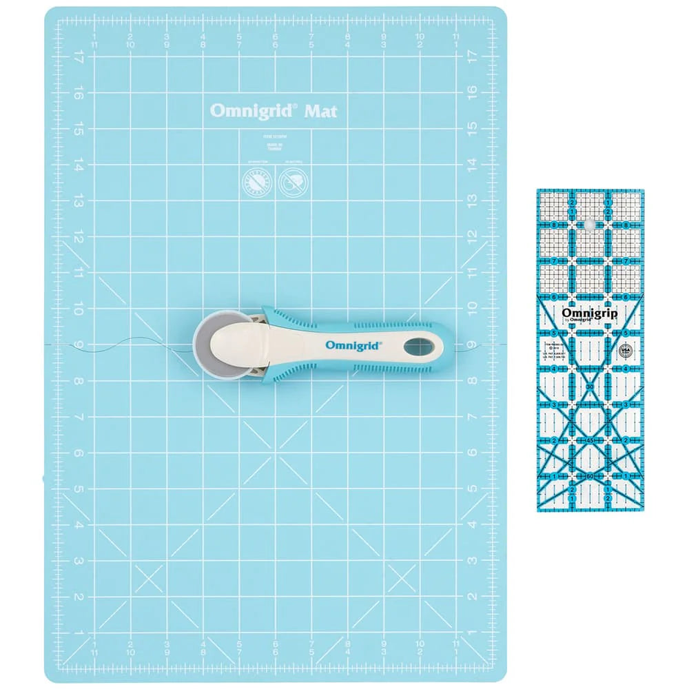 The Tacony Corporation Omnigrid 12 x 18 Compact Rotary Cutting Kit, featuring a blue cutting mat, ergonomic blue-handled rotary cutter, and clear quilting ruler, is displayed side by side on a white background.