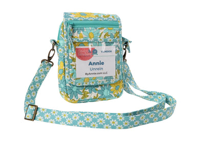 A small quilted crossbody bag made with the On the Go 2.0 Quilt Pattern, featuring a blue/yellow floral design, ByAnnie.com LLC name badge, front zipper pocket, and adjustable strap.