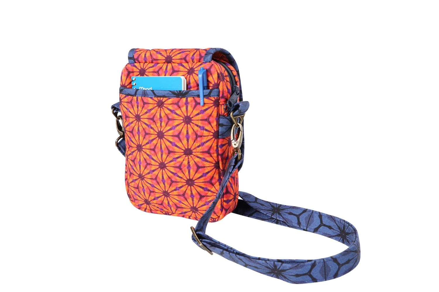A small crossbody bag in orange and purple geometric floral print, made using the On the Go 2.0 Quilt Pattern. It has a front pocket holding a blue card and a matching adjustable strap, displayed on a white background.