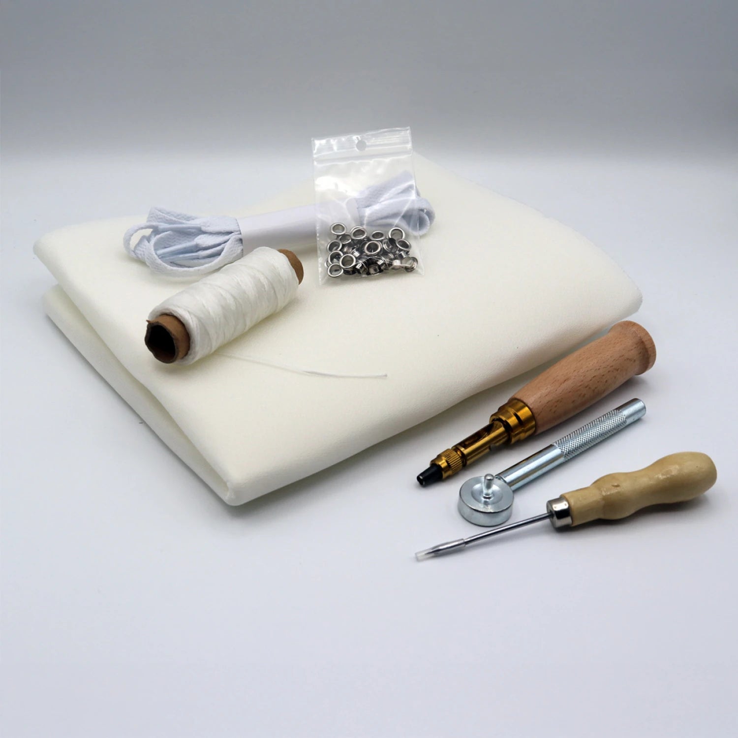 The Patchwork Kicks White Shoe Kit by Checker Distributor includes fabric foam, white thread, cord, metal grommets in a bag, a wooden-handled awl, screwdriver, and metal tool—perfect for beginner sewing or sneaker-making kits.