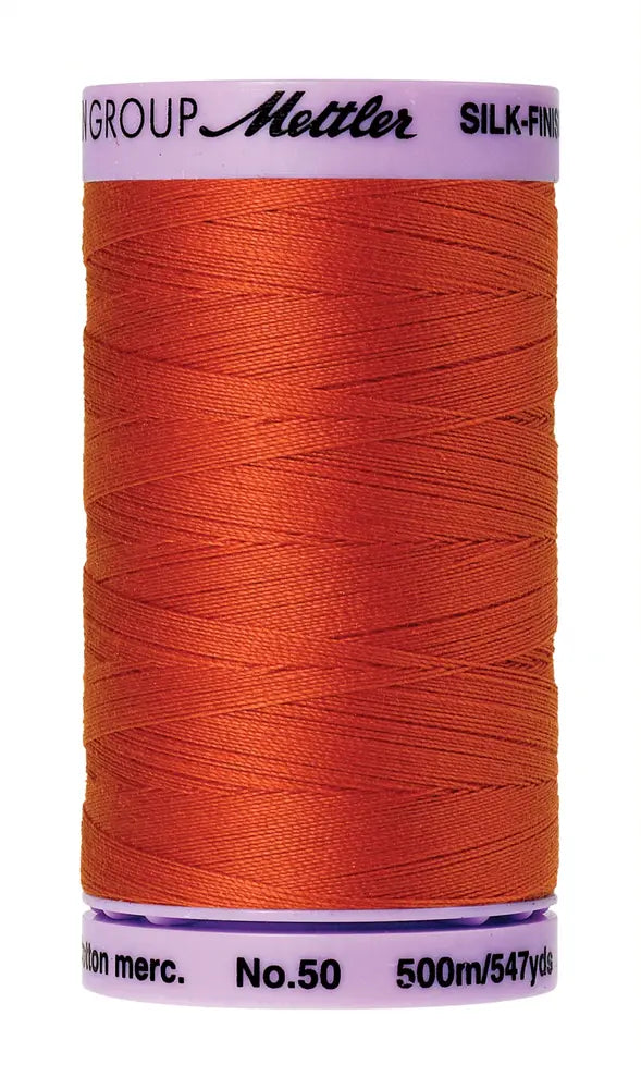 A spool of Mettler Cotton Silk Finish 50wt 500m quilting thread in Paprika with a white cap featuring brand and product details.