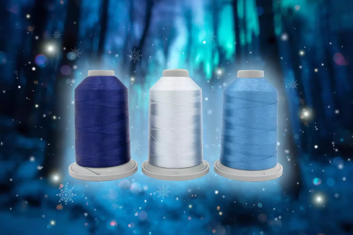 Three spools from the Hab+Dash Polar Prism Glide Thread of the Month Bundle in dark blue, white, and light blue are displayed side by side against a sparkling, wintry blue background with snowflake patterns.