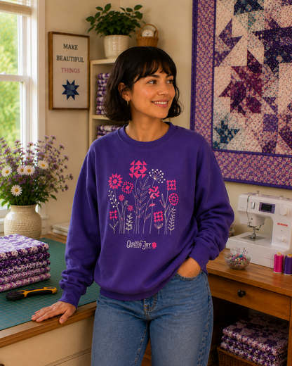 A woman in a Quilted Joy Flowers Indigo Crewneck Sweatshirt stands smiling in a cozy sewing room with quilts and flowers—a thoughtful gift for quilters from Quilted Joy.