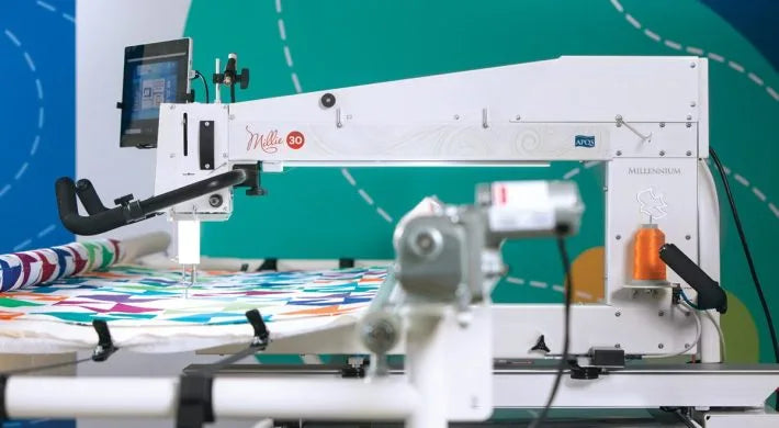 The APQS Quilt Path Robot Computer by APQS features a touchscreen display and thread spool, shown quilting a vibrant quilt against a bright blue and green background.