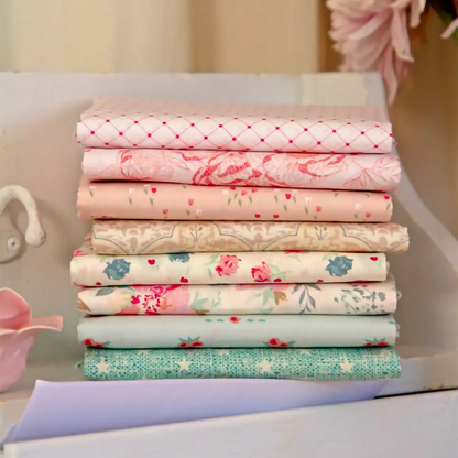 A stack of eight pastel fabrics from the Romantica Half Yard Bundle, featuring floral and vintage-inspired quilting patterns, is neatly arranged on a white wooden surface with soft decor in the background.