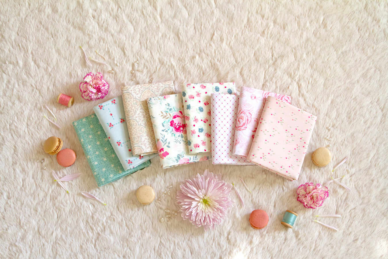 Seven pastel fabric squares from the Romantica Half Yard Bundle, featuring floral and geometric prints, are arranged on a cream surface. Pink flowers, macarons, and petals create a soft, cozy atmosphere.