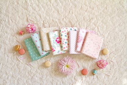 Seven pastel fabric squares from the Romantica Half Yard Bundle, featuring floral and geometric prints, are arranged on a cream surface. Pink flowers, macarons, and petals create a soft, cozy atmosphere.