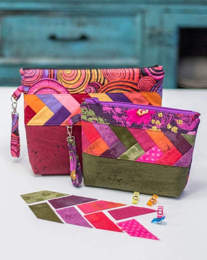 Two vibrant quilted zipper pouches made with geometric patterns in pink, purple, orange, and green are shown on a white surface. The Pack it Up Pattern by Checker Distributor is featured with fabric swatches and sewing clips in front.