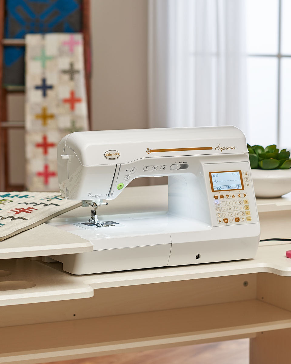 A computerized sewing machine sits on a table with a quilting project under the needle; a quilt with colorful cross patterns hangs in the background and a small plant is nearby.