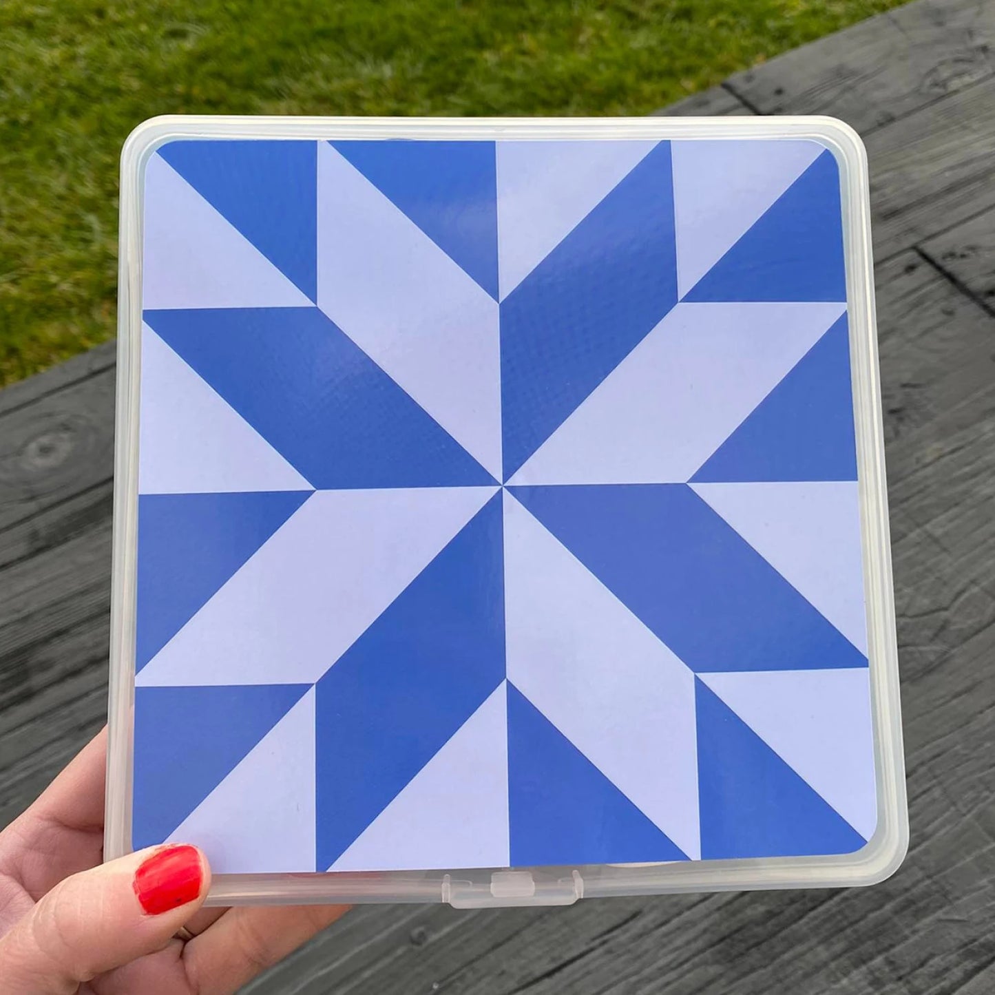 A hand with red nail polish holds the Checker Distributor Summit Street Storage Box - Bluebird, showcasing its blue and white geometric star pattern, with a green lawn and wooden deck in the background.