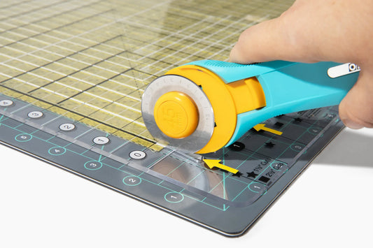 A hand uses a turquoise rotary cutter to cut fabric on a gridded mat, guided by transparent ruler guides and the Quilted Joy Stripology ruler—just like in "Beyond the Strip: Mastering Your Stripology Ruler.