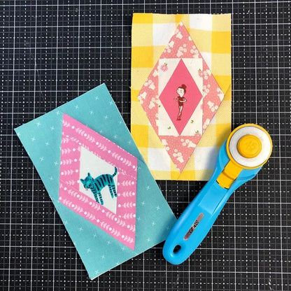 Two quilt blocks—one with a girl on yellow plaid, one with a blue cat on teal—made using the Scrappy Diamonds Foundation Pad by Checker Distributor, are placed on a black grid background next to a blue and yellow rotary cutter.