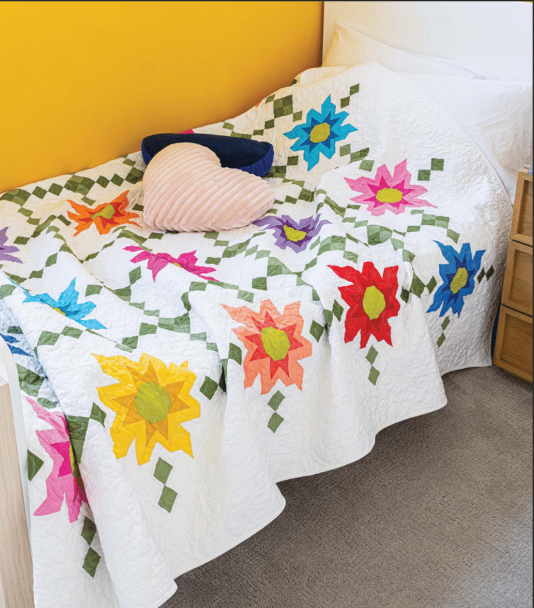 A neatly made bed showcases the Quilted Joy Love of Quilting Wildflower Blossoms Quilt Kit, with bright flowers and green leaves on white. A heart-shaped pink pillow and a dark blue round pillow rest on top.