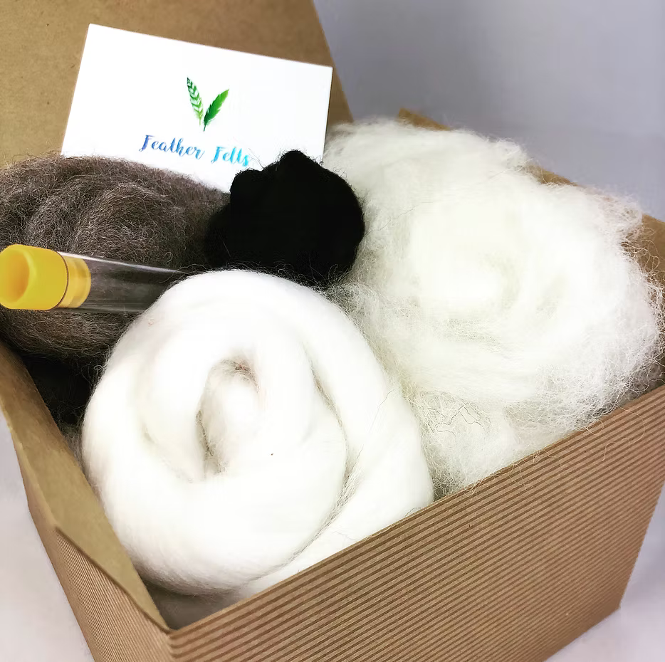 The Grey Sheep - Needle Felting Kit No Foam by Feather Felts includes rolled white wool, a dark brown ball, a small black bundle, a clear tube, and a Feather Felts business card—ideal for beginner needle felting projects.