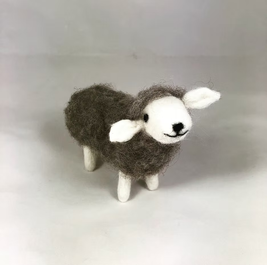 The Feather Felts Grey Sheep - Needle Felting Kit No Foam lets you create a handmade felt sheep with a dark gray body and white legs, face, and ears. The finished sheep stands on a plain white surface.