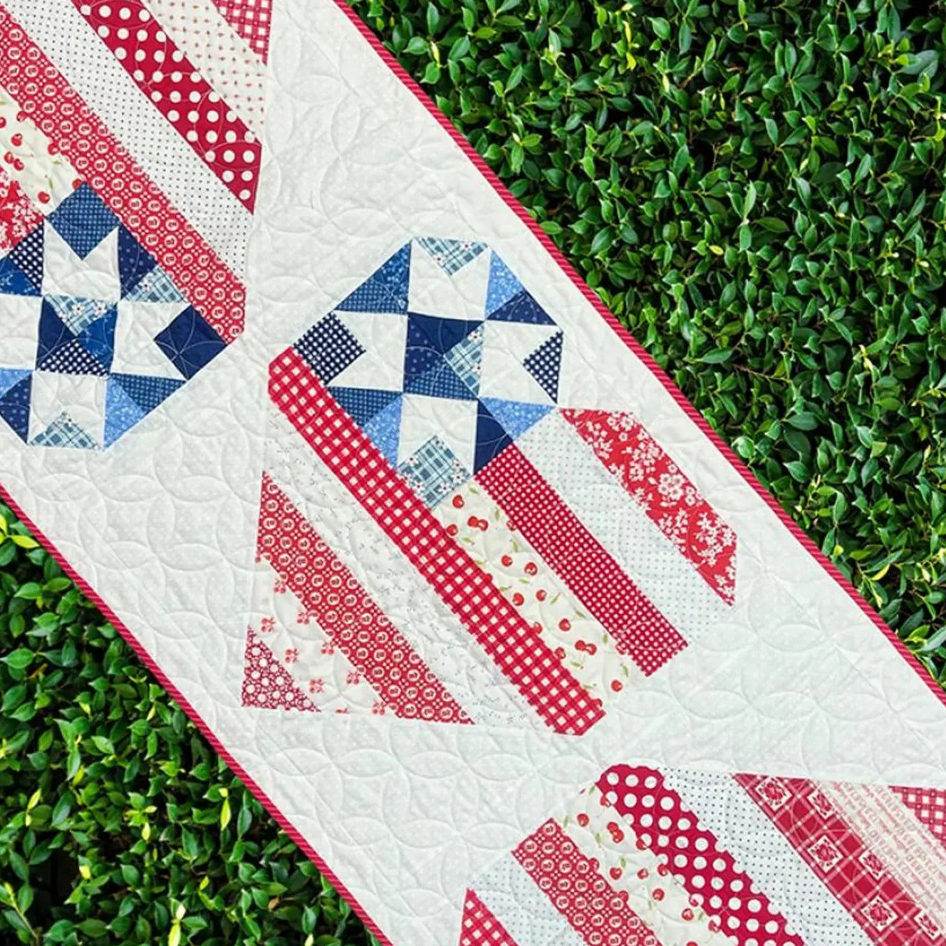 The Star Spangled Table Runner by Quilted Joy features red, white, and blue geometric star patterns with striped sections, giving your table a festive Americana look. It’s shown diagonally atop green leafy ground for a patriotic display.