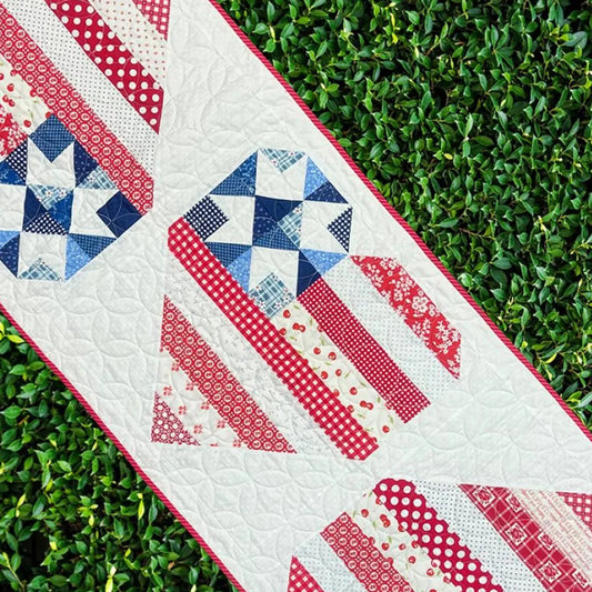 The Star Spangled Table Runner by Quilted Joy features red, white, and blue geometric star patterns with striped sections, giving your table a festive Americana look. It’s shown diagonally atop green leafy ground for a patriotic display.