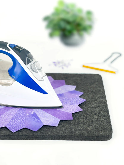 A clothes iron presses a purple, flower-shaped quilt piece on the Checker Distributor Wool Pressing Mat 17in x 24in x 1/2in Thick. In the blurred background are a green potted plant, sewing pins, and a yellow-handled seam ripper.