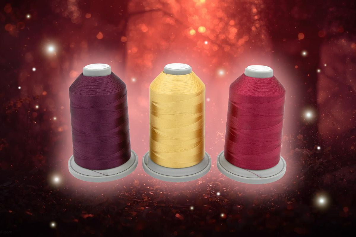 Three spools of thread—burgundy, yellow, and red—from the Velvet Vineyard Glide Thread of the Month Bundle by Hab+Dash by Fil Tec are displayed side by side against a glowing, warm-toned background.
