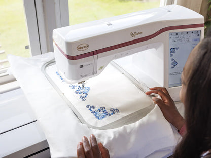 A person uses the Baby Lock Reflection Sewing & Embroidery machine with FREE 60 Days of Online Classes to stitch a blue floral pattern on white fabric; its digital screen displays the design near a bright window.