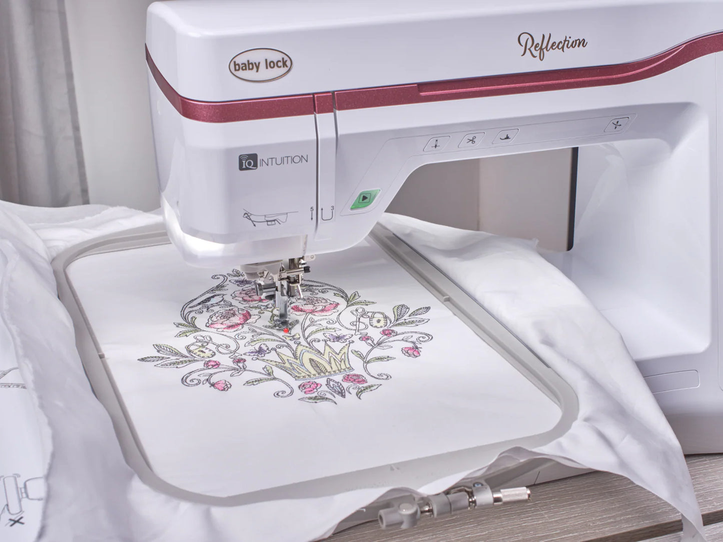 A Baby Lock Reflection Sewing & Embroidery machine, with FREE 60 Days of Online Classes, stitches a detailed floral and crown design onto white fabric in an embroidery hoop, mid-operation on a worktable.