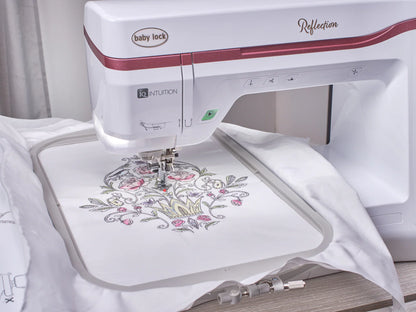 A Baby Lock Reflection Sewing & Embroidery machine, with FREE 60 Days of Online Classes, stitches a detailed floral and crown design onto white fabric in an embroidery hoop, mid-operation on a worktable.
