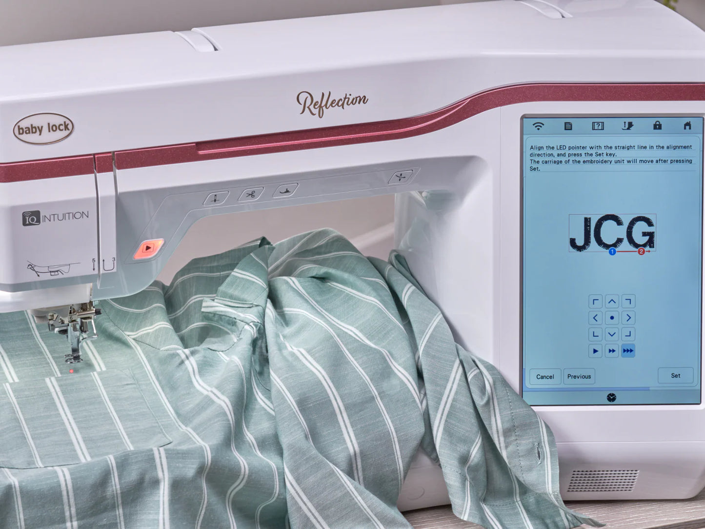 The Baby Lock Reflection Sewing & Embroidery machine stitches a green and white striped fabric, displaying blue and red "JCG" initials on its large screen. This model includes FREE 60 Days of Online Classes.