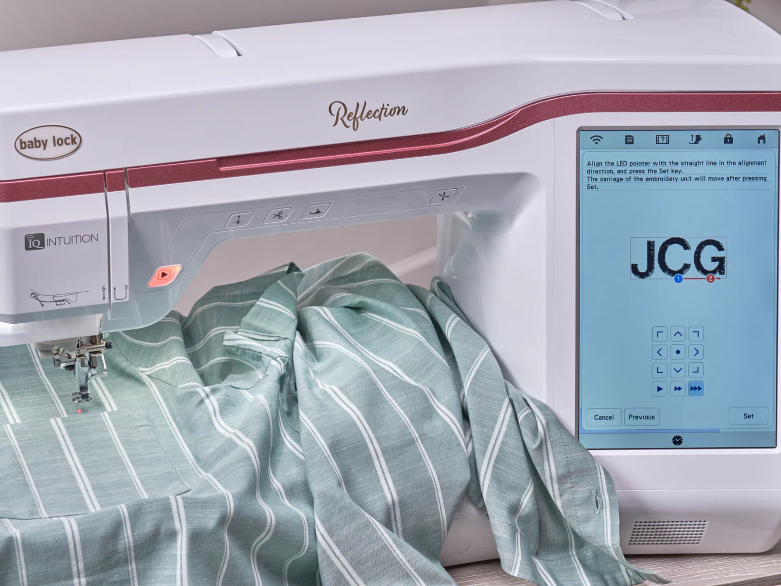 The Baby Lock Reflection Sewing & Embroidery machine stitches a green and white striped fabric, displaying blue and red "JCG" initials on its large screen. This model includes FREE 60 Days of Online Classes.