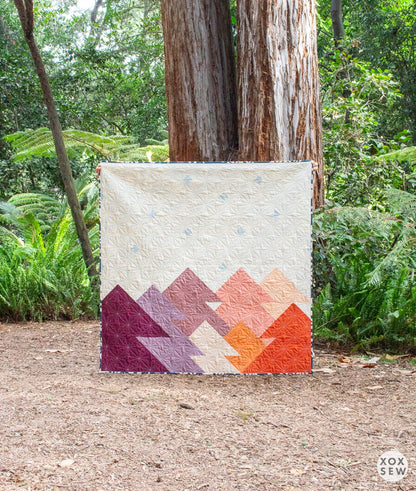 The Forest Quilt Pattern by Checker Distributor features a modern geometric mountain design in purple, pink, and orange—perfect for showcasing outdoors amid tall trees and lush greenery.