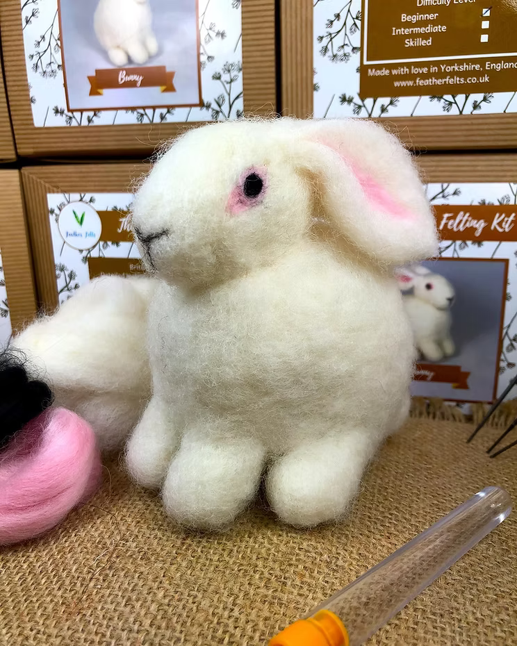 A handmade white wool felt bunny by Feather Felts sits on burlap, with Bunny - Needle Felting Kit No Foam boxes behind it. A felting needle and wool are in the foreground, matching the kit’s bunny design.