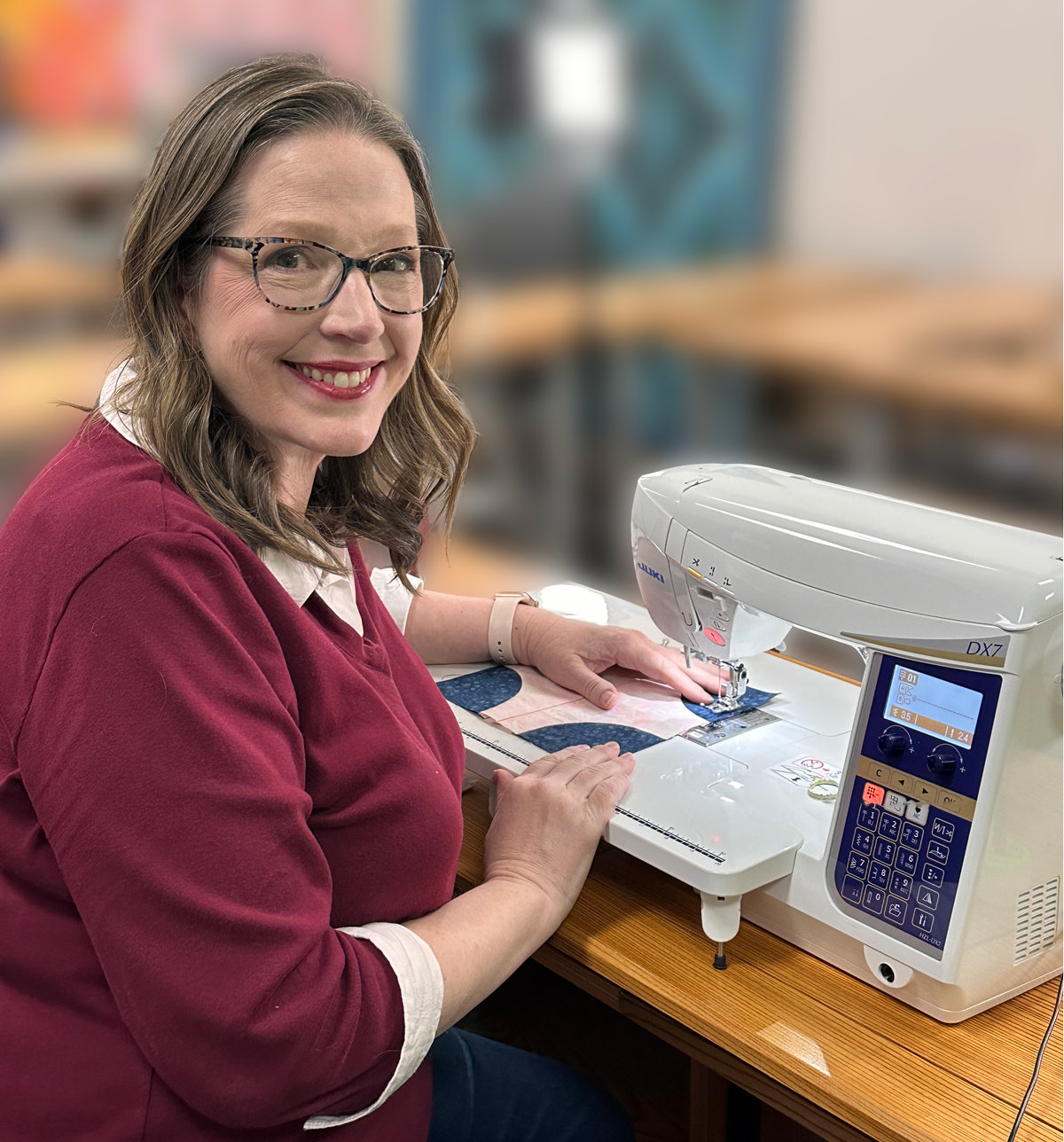 A woman wearing glasses and a maroon sweater smiles while using a sewing machine at a wooden table in a bright, blurred classroom or workshop setting.
