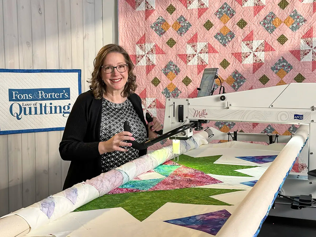 A smiling woman at the APQS Roadshow by Quilted Joy works on a vibrant quilt with a longarm quilting machine, standing before a quilted backdrop and a Fons & Porter’s Love of Quilting sign—highlighting her successful quilting business.