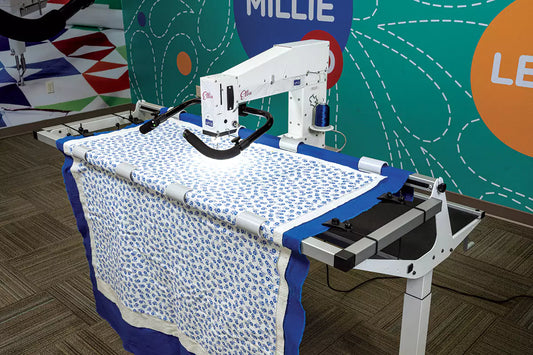 An APQS Ellie 18" Long Arm Quilting Machine with FREE Online Class quilts a blue and white patterned blanket in a vibrant room adorned with abstract wall art, while the word MILLIE appears in the background.