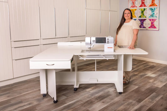 A woman stands beside the Arrow Christa Sewing and Quilting Machine Cabinet by Arrow Companies, LLC in a bright room with wood floors, built-in cabinets, and a colorful quilt displayed on the wall.