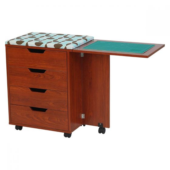 The Arrow Laverne and Shirley Sewing and Quilting Machine Cabinet by Arrow Companies, LLC features four drawers, a geometric print cushioned seat, a fold-out side table with green cutting mat, and black wheels for easy mobility.