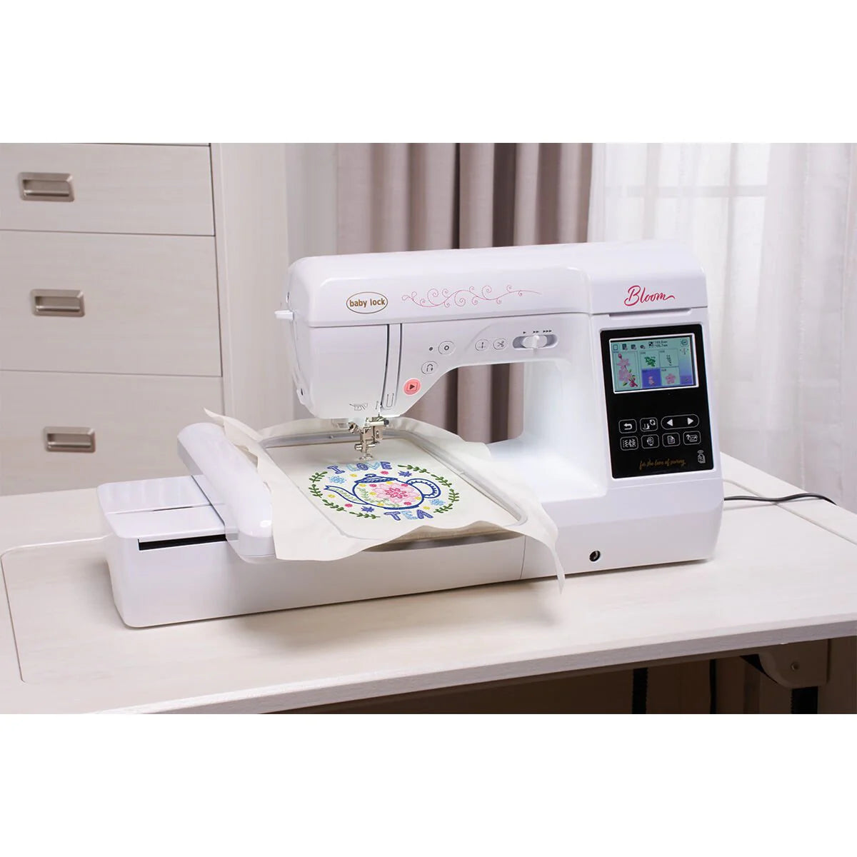 A Baby Lock Bloom Sewing & Embroidery Machine by Baby Lock stitches a vibrant floral design on white fabric, its digital display showing the pattern while set on a light desk in a bright room with drawers and curtains.