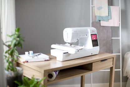 The Baby Lock Bloom Sewing & Embroidery Machine by Baby Lock rests on a wooden desk with notebooks, thread spools, and a pen nearby. A plant sits to the left, while folded fabrics are neatly arranged in the background on a white rack.