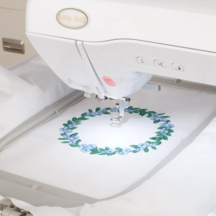 The Baby Lock Flare Dedicated Embroidery Machine with FREE Online Classes is stitching a circular floral wreath with blue flowers and green leaves onto white fabric, showing its precision for intricate designs and embroidery fonts.