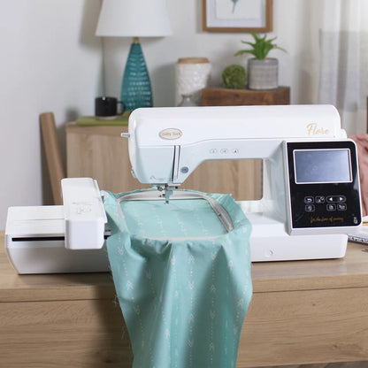 A Baby Lock Flare Dedicated Embroidery Machine with FREE Online Classes is on a wooden table, stitching teal fabric with an arrow pattern. Decor, plants, and home items are visible in the background.